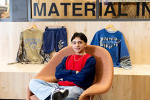 A person sits in a modern chair with arms crossed, with three graphic clothing items displayed on the wall behind them.