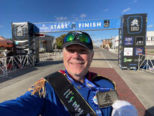 A man wearing a blue Prince Charming-style costume takes a selfie while holding his marathon finisher medal. Behind him is the Crossroads Marathon start/finish archway on a sunny day.
