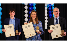 Three award recipients pose with certificates and medallions in front of blue and white balloon columns.