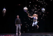 A performer jumps midair as confetti bursts around them on a dark stage, while another performer stands nearby watching; three hanging props resembling piñatas or suspended sculptures are positioned above them.