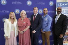 Five people stand in front of a blue Commonwealth of Kentucky-branded backdrop during an event, with a vertical display banner visible at the right side of the photo.