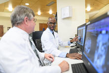 Two medical professionals in white lab coats sit at computer workstations in a clinical setting, reviewing medical imaging on large monitors as they discuss the results.