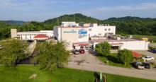 Aerial view of a large light-colored medical building complex with multiple entrances, parked vehicles and ambulance bays, surrounded by green trees and rolling hills under a clear sky.