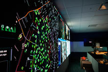 A darkened control room with a wall-sized digital map displaying green data points and lines, surrounded by monitors and workstations.