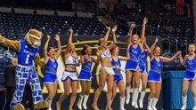 University mascot and dance team perform on a basketball court, raising their arms during a routine in an arena.