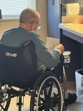 An older man in a wheelchair reads a handwritten letter near a counter in a bright room. He wears glasses and a long-sleeve shirt. A cup is attached to his wheelchair armrest, and light comes in through a nearby window.