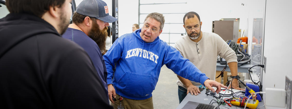 Group of people gathered around a workbench, looking at electronics and tools in a workshop.