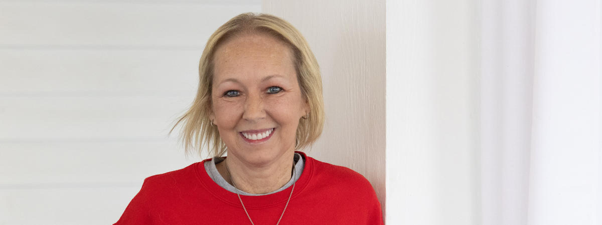 Vikki Lane smiles at the camera while standing in front of a white wall. She is wearing a red sweater with a gray collar and a silver necklace.