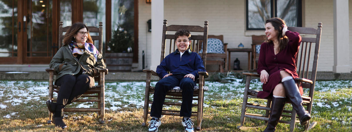 Three people sitting on wooden rocking chairs in a grassy yard with light snow, in front of a house with a covered porch.
