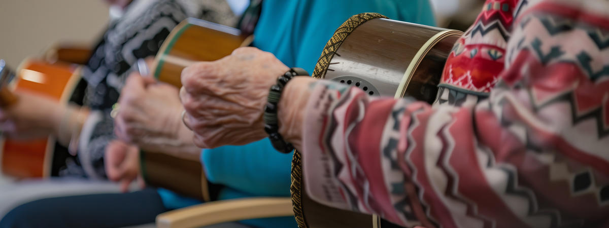 Three older adults sit side by side, playing hand drums, as part of a small group music session or recreational activity in a community or senior center.