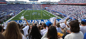 Fans fill a packed stadium as players line up on the field at a University of Kentucky football game, with the crowd waving white rally towels.