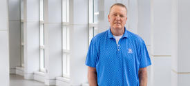 A person stands in a bright, modern hallway with tall windows and white architectural panels. They are wearing a blue short‑sleeve polo shirt with a subtle dotted pattern and khaki pants