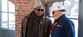 Two people stand indoors near a brick wall and windows, talking. One wears a knit cap and jacket; the other wears a hard hat and holds a clipboard.