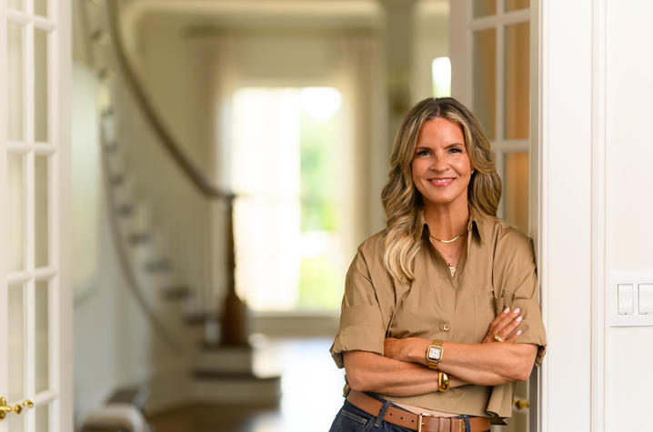 A person stands with arms crossed in the doorway of a bright residential-style interior. The person is wearing a short-sleeve tan button-up shirt, jeans and a brown belt. The background features white walls, French doors and a curved staircase.