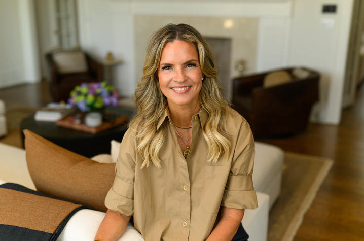 A person wearing a tan button-up shirt sits in a bright living room with hardwood floors and neutral-colored furnishings. The room features a fireplace, floral arrangement, upholstered chairs and a seating area in the background.