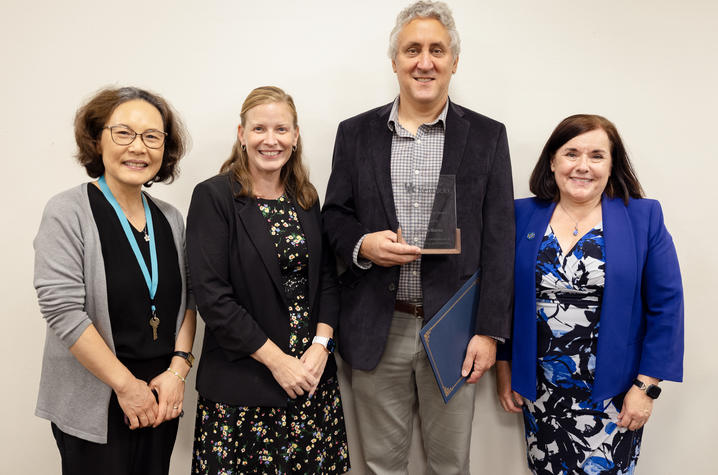 Four people in a group photo, with one person holding a glass award and a blue certificate folder against a plain indoor background.