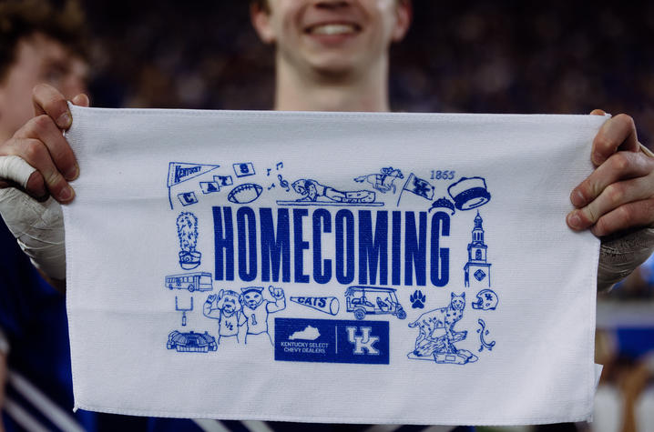 Close-up of a person holding a white rally towel featuring a blue “Homecoming” design with University of Kentucky branding and illustrated campus-themed icons. A blurred crowd and another person are visible in the background.