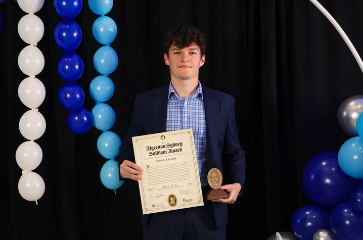 A person in a blue suit stands in front of blue and white balloon decorations, holding a framed certificate and a medallion award.