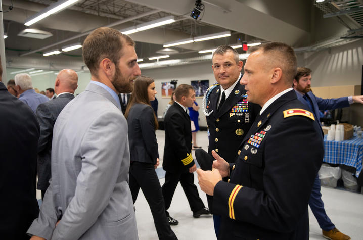 Three men stand in conversation at an indoor event. Two men in U.S. Army dress uniforms with ribbons and insignia speak with a man in a light gray suit. 