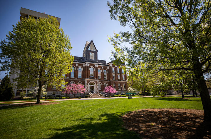 The front of Main Building at the University of Kentucky during the springtime, with trees and grass in the foreground and a clear blue sky in the background. 