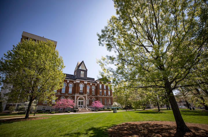 Historic red-brick campus building with arched windows and a central tower, framed by large green trees and a wide lawn, with pink flowering trees in bloom along the walkway on a sunny spring day.