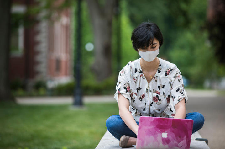 student sitting on wall working on computer and wearing mask