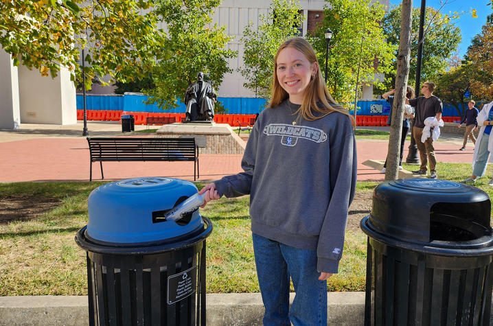 A smiling female student stands next to a recycling can. 