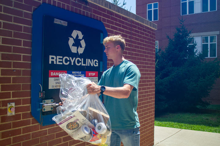 A male student in a t-shirt and jeans places materials to be recycled into a recycling container. 