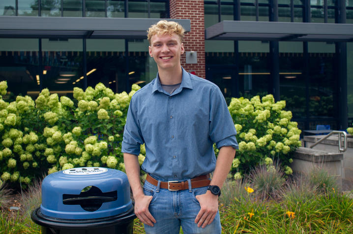 A smiling male student stands next to a campus recyling can. 