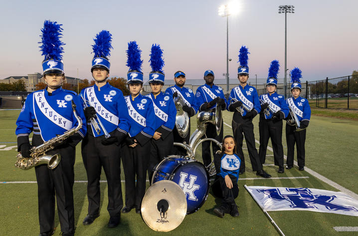 A group of nine members of the UK marching band pose for a photo on a football practice field.