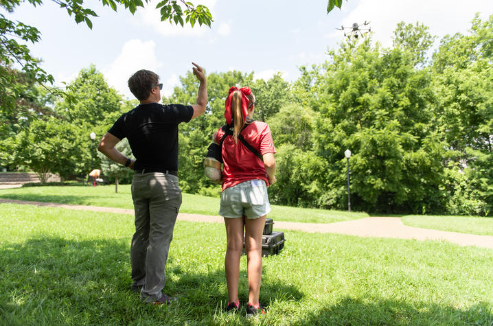 An instructor and a youth stand in a grassy park, watching a drone fly overhead. The instructor points toward the aircraft while the student, wearing a red shirt and bow, observes a hands-on drone operation activity.