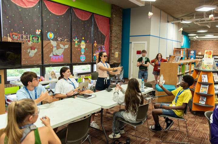 An instructor leads a group of students gathered around tables in a library classroom. Participants engage in a hands-on activity while discussing ideas, with colorful circus-themed artwork decorating the windows behind them.