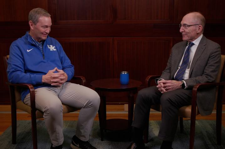 Two people sit across from each other in chairs on either side of a small round table in a wood‑paneled room. One wears a UK athletic-style jacket and light-colored pants, while the other wears a suit and tie. A small blue object sits on the table between