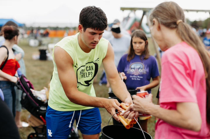 A volunteer wearing a neon green “How Can I Help?” shirt assists participants at an outdoor community event. Several people stand nearby, and activity stations are visible in the background.