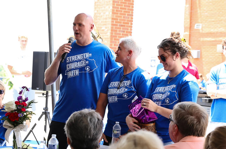 Three people wearing matching blue event T-shirts stand before a seated audience while one person speaks into a microphone. Tables with flowers and water bottles are visible in the foreground.