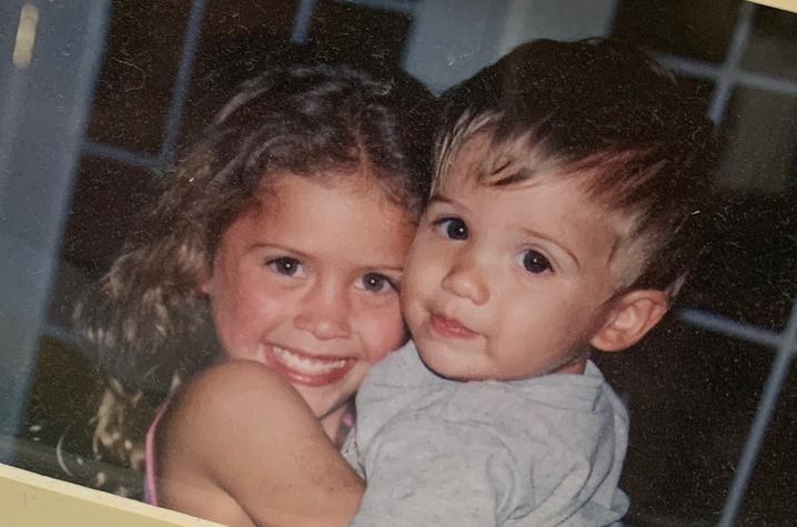 A close-up childhood photograph shows two young children embracing and looking toward the camera indoors. One child is holding the other, and both are prominently framed against a softly blurred background.