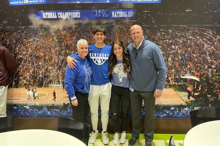 Four people pose together in front of a large mural depicting a University of Kentucky basketball championship celebration. The group stands with their arms around one another, wearing Kentucky-themed clothing, confetti and a packed arena crowd in backgro