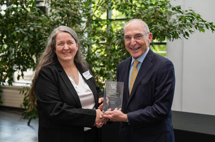 Two people shake hands while holding a glass award during a recognition ceremony. They stand in a bright indoor space with large leafy plants behind them, celebrating an academic achievement.