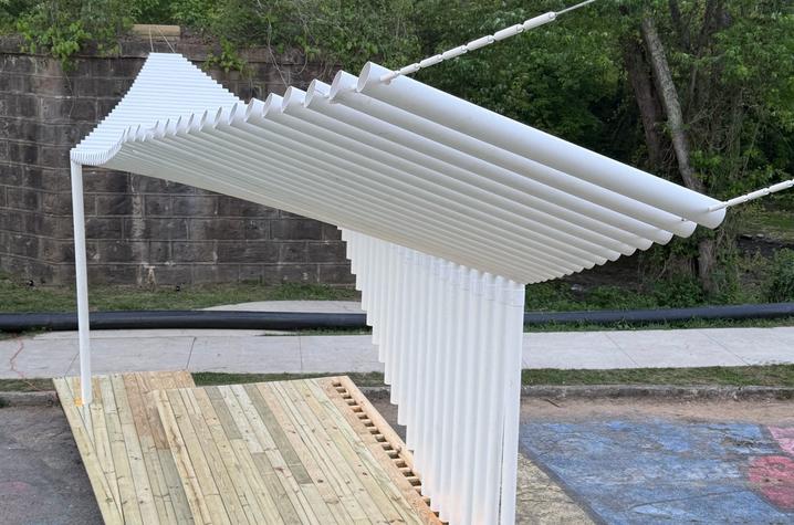 A modern white canopy made of suspended cylindrical tubes extends over a wooden deck and walkway. The sculptural structure sits in an outdoor space surrounded by trees, with a stone retaining wall and colorful painted pavement visible below.