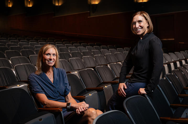 Two people pose among rows of seats in a dimly lit auditorium, with one seated and the other leaning against a nearby chair. Wall lights illuminate the theater in the background.