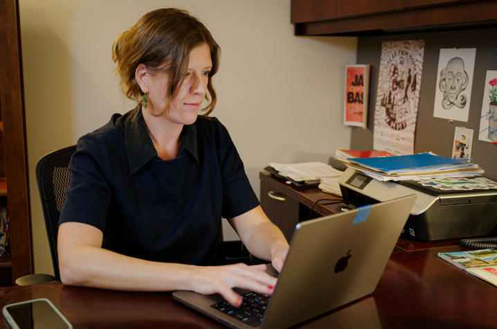 Person seated at a desk using a laptop in an office workspace with books, papers and artwork visible in the background.