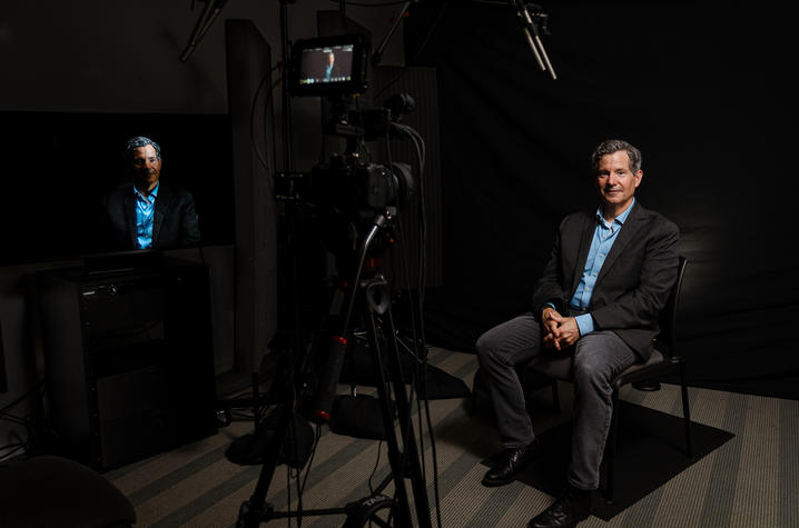 A person sits in a recording studio during a video interview. A camera, lighting equipment and microphones are visible in the foreground, capturing the subject against a dark backdrop while a monitor displays the live video feed.