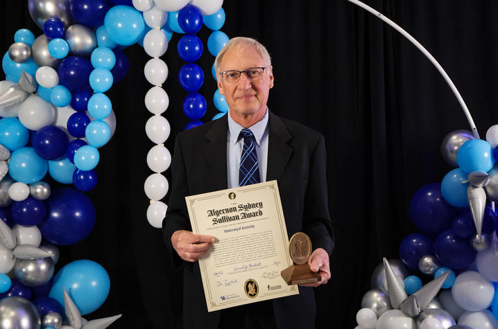 An individual stands in front of blue, white and silver balloon decorations, holding a framed certificate and a medallion award.
