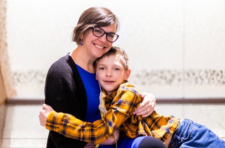 Image of Dynae Utz in blue dress with black sweater, hugging her nine-year-old son Lewis who is wearing a yellow plaid shirt and jeans.