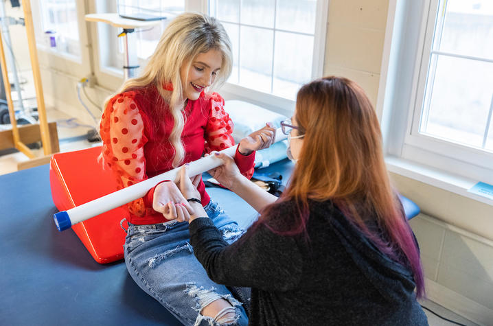 kayla sitting, working with an occupational therapist. she's working on her arm and core strength, using a pole to pull herself up.