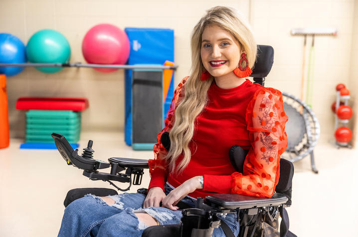 kayla lacy wearing a very cute red blouse, sitting in her wheelchair within the therapy clinic, smiling for the camera.