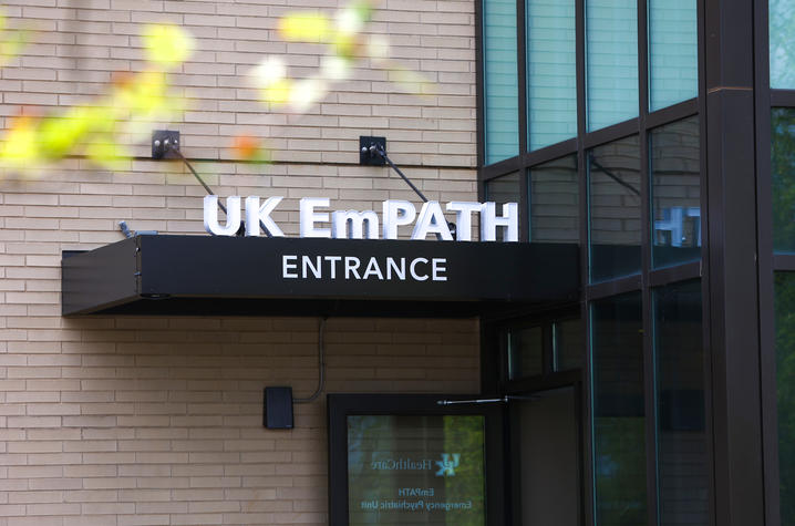 Photo of the EmPATH entrance Sign reading “UK EmPATH Entrance” above a glass doorway on a brick building, with soft green leaves blurred in the foreground.