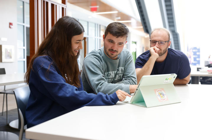Photo of pharmacy students Three students sit together at a table, collaborating while looking at a tablet in a bright, modern campus building. One student points at the screen as the others lean in, focused and engaged in discussion.