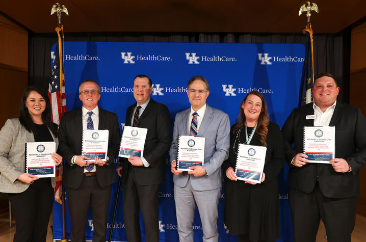 A group of six individuals is standing in front of a blue backdrop with the text “UK HealthCare” and the UK logo repeated across it. They are holding certificates or award booklets. Two American flags are positioned on either side of the backdrop.