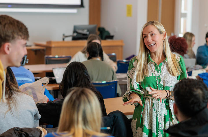 An instructor wearing a green dress talks to a group of students in a classroom.